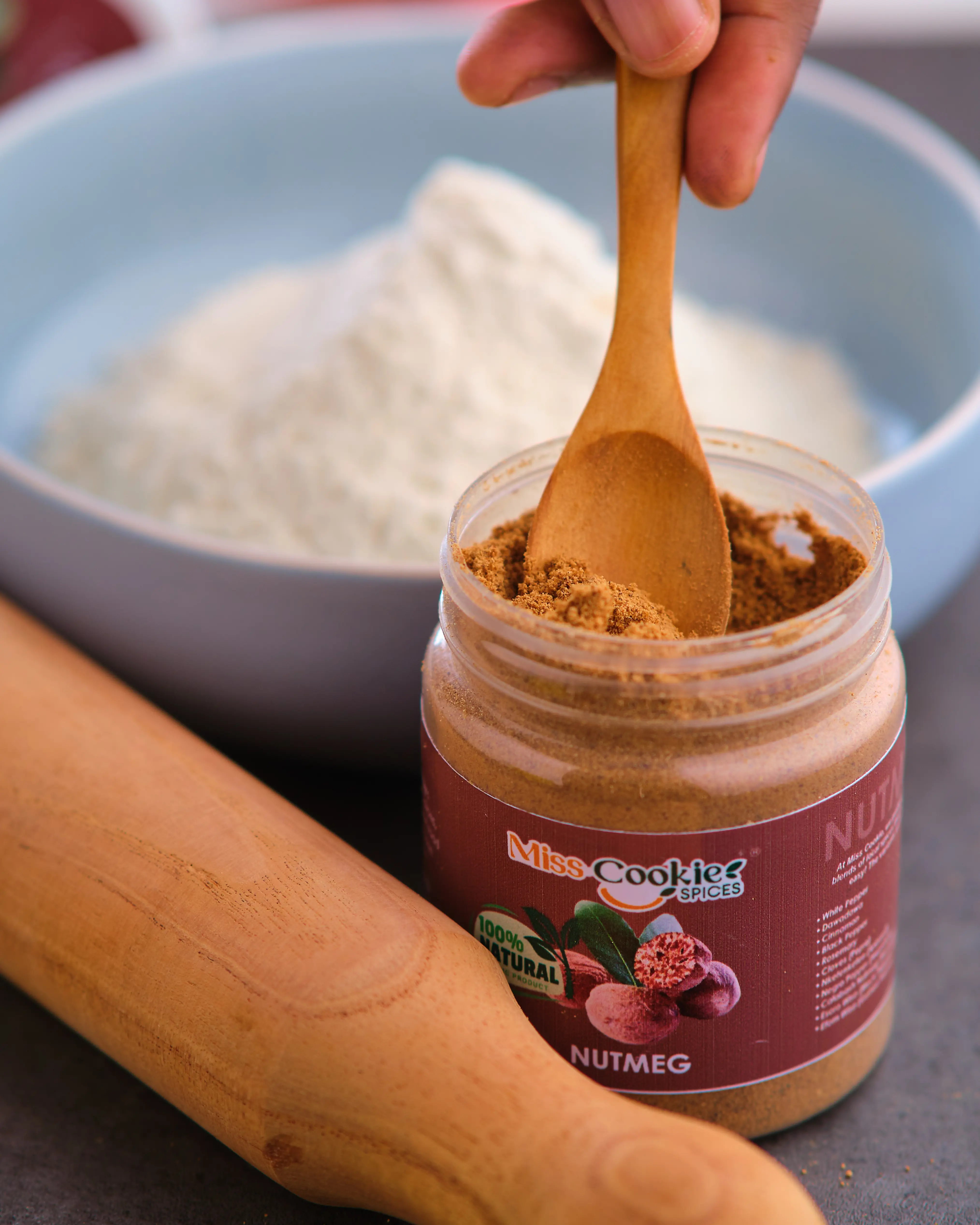Wooden spoon scooping Miss Cookie nutmeg beside a bowl of flour and a rolling pin